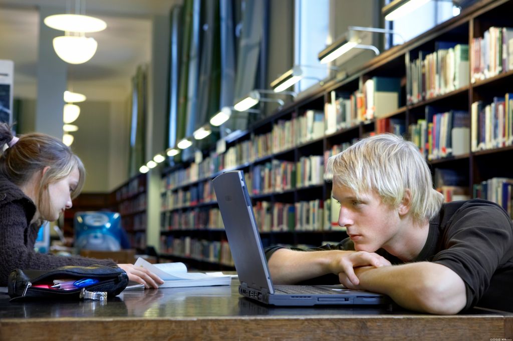 To elever sitter inne på et bibliotek og jobber konsentrert. Den ene eleven sitter bøyd over pc-en med haka på kryssede armer. Den andre eleven leser i ei bok. Foto.