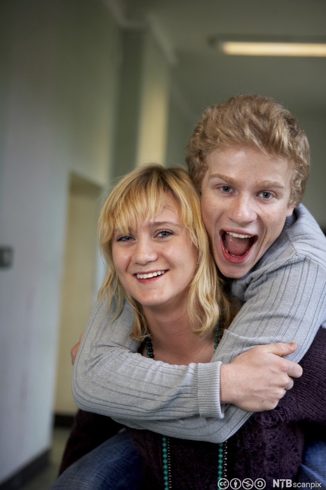 Gutt omfavner jente bakfra i en skolekorridor. Begge smiler og ler. Foto. 