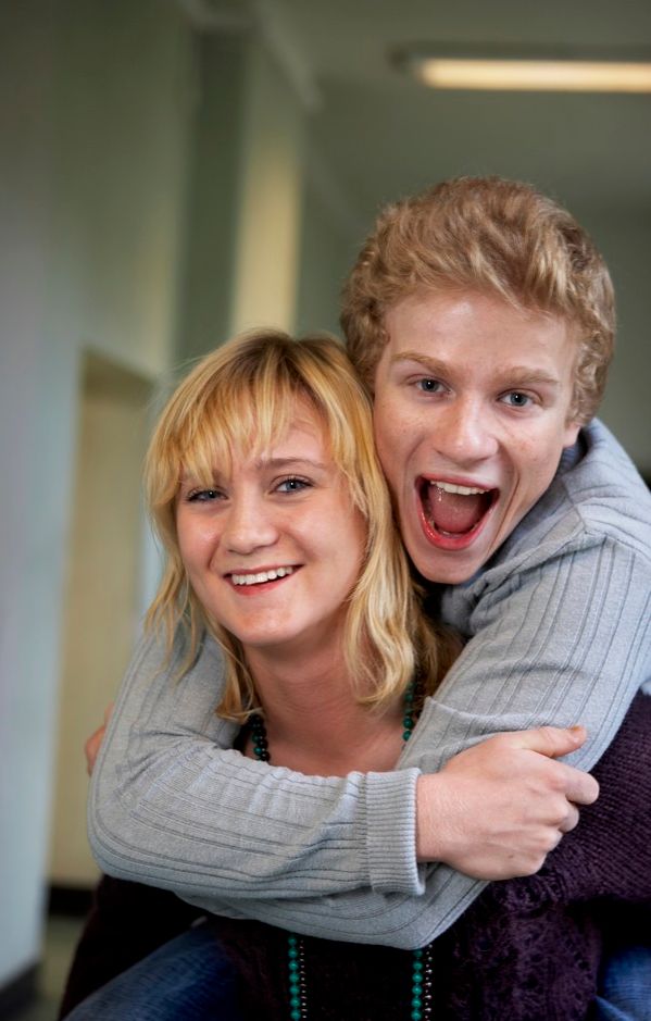 Gutt omfavner jente bakfra i en skolekorridor. Begge smiler og ler. Foto.