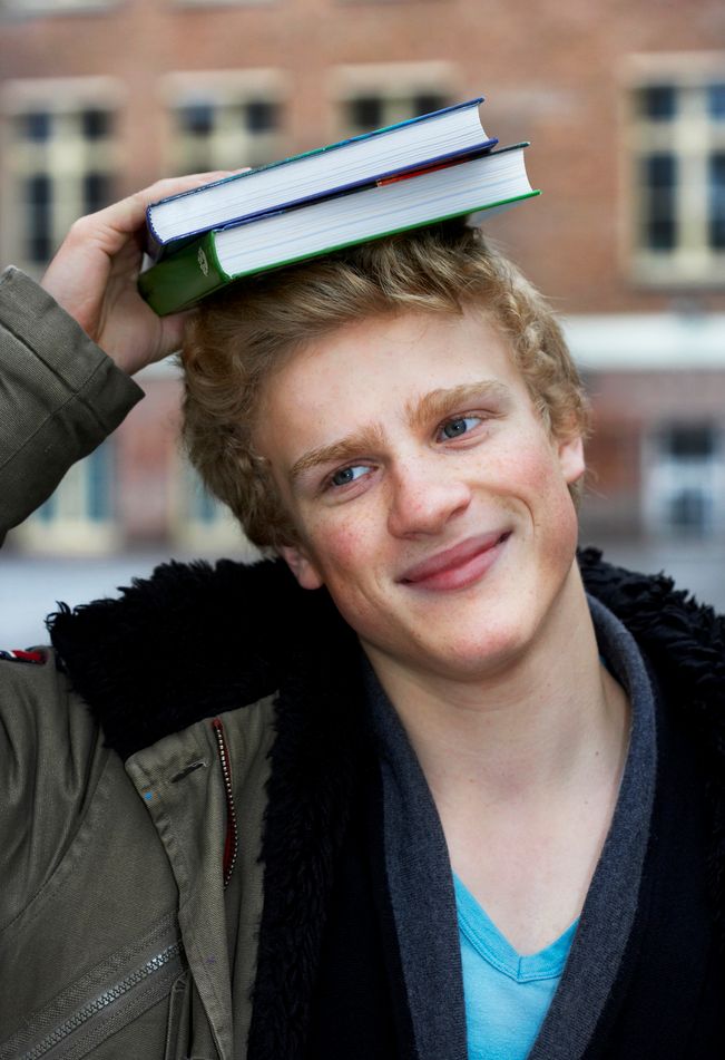 En gutt står utenfor et skolebygg. Han holder to bøker på hodet og smiler. Foto.