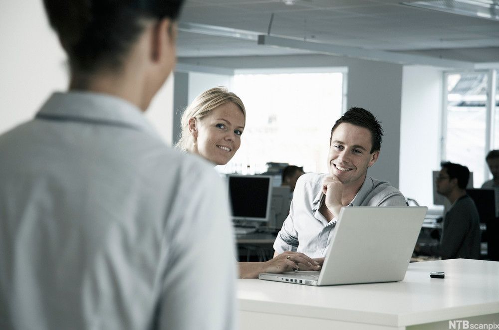 En kvinne sitter ved en laptop, en mann sitter ved siden av. Begge ser mot en tredje person som nærmer seg. Foto.