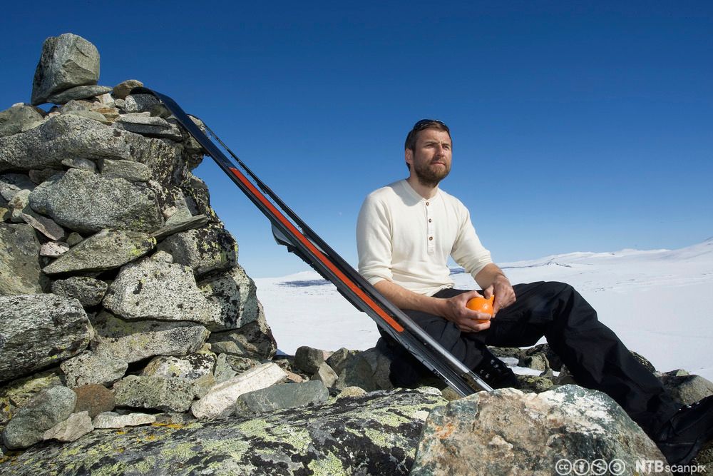 Mann spiser en appelsin på en fjelltopp. Et par ski er stilt opp mot en varde ved siden av ham. Foto.