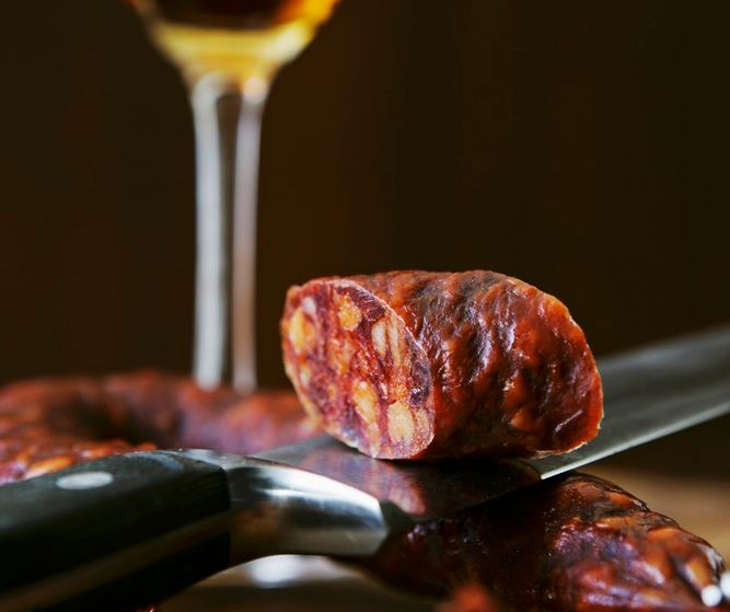 Et stykke chorizopølse på en kniv og et glass sherry i bakgrunnen. Foto.
