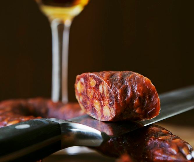 Eit stykke chorizopølse på ein kniv og eit glas sherry i bakgrunnen. Foto.