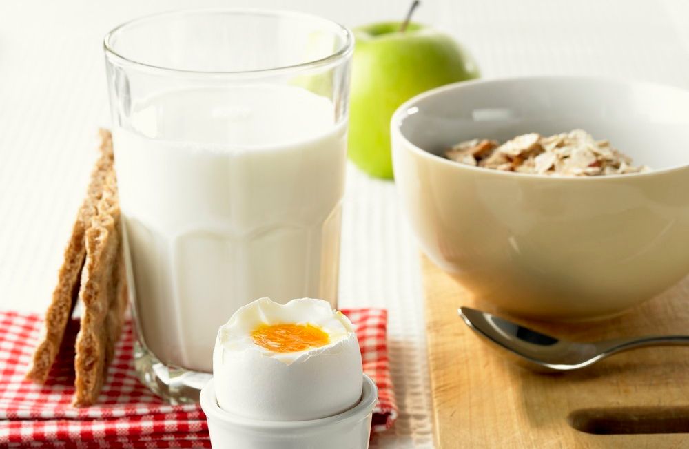 Foto av et måltid bestående av bløtkokt egg i et eggeglass, et glass melk, en bolle med frokostblanding, et par knekkebrød og et eple.
