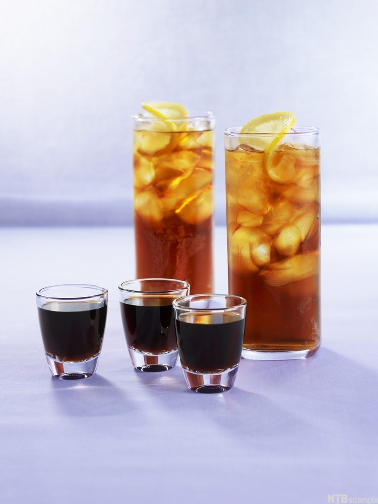 Tre snapsglass med svart væske og to drinkglass med lys brun væske, isbiter og sitronskive på toppen. Foto.