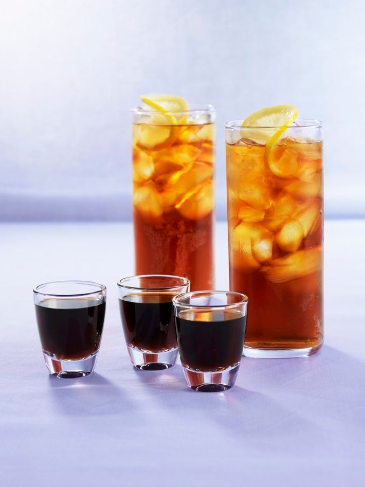 Tre snapsglass med svart væske og to drinkglas med lys brun væske, isbitar og sitronskive på toppen. Foto.