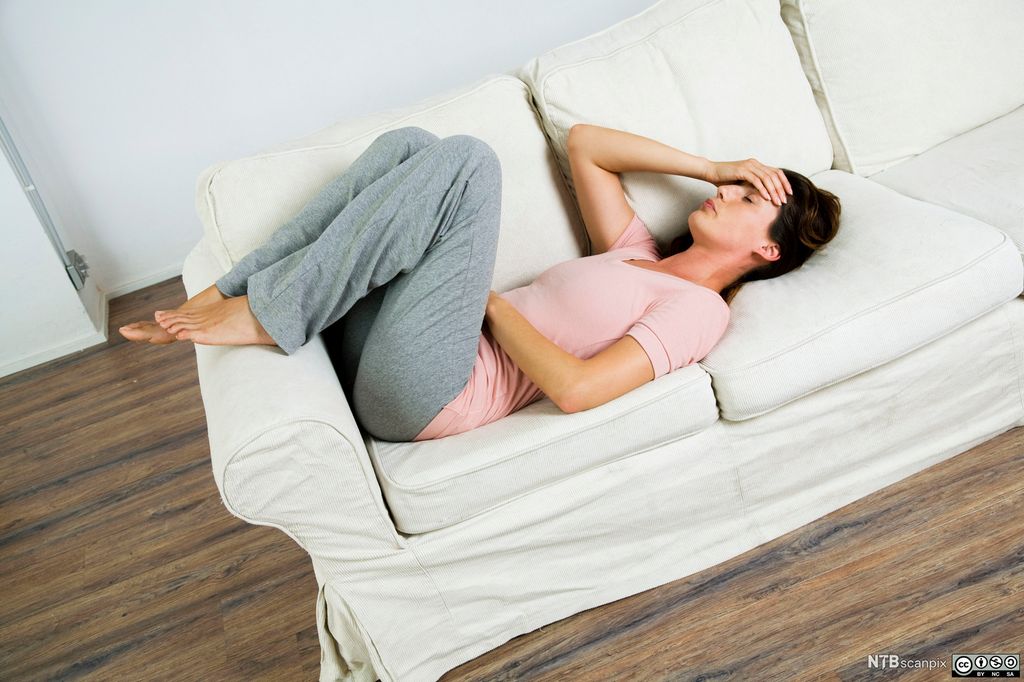 Kvinne ligger på sofa med ei hånd på hodet og ei hånd på magen. Foto.