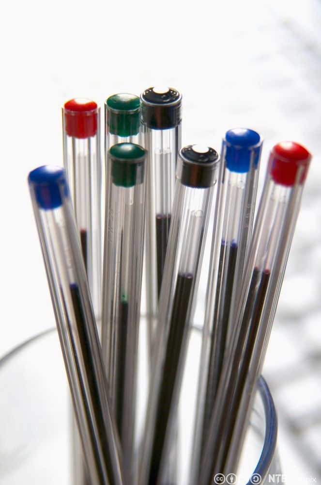 Åtte gjenomsiktige kulepennar i plast stikk opp av eit glas. På toppen er det eit plastlokk som viser fargen på blekket: svart, raudt, grønt eller blått. Foto.