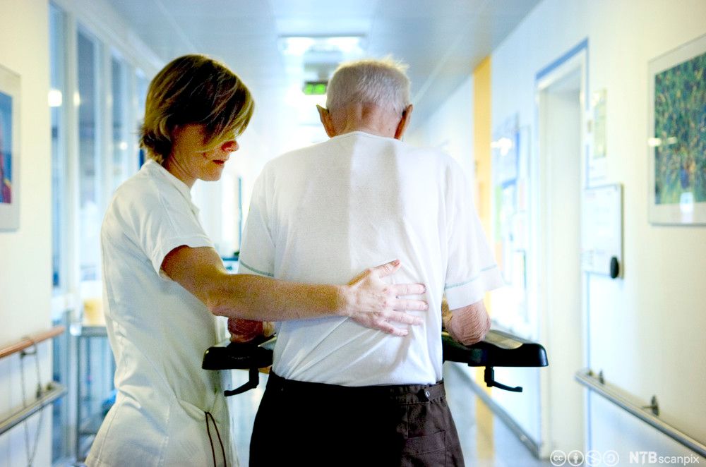 Helsefagarbeider hjelper eldre mann med gåbord i en korridor på et sykehus eller et sykehjem. Foto.