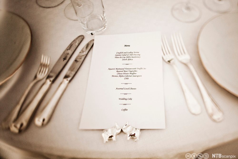 En kuvert på et bord med hvit duk dekket til selskap med en meny i midten mellom bestikket. Foto.