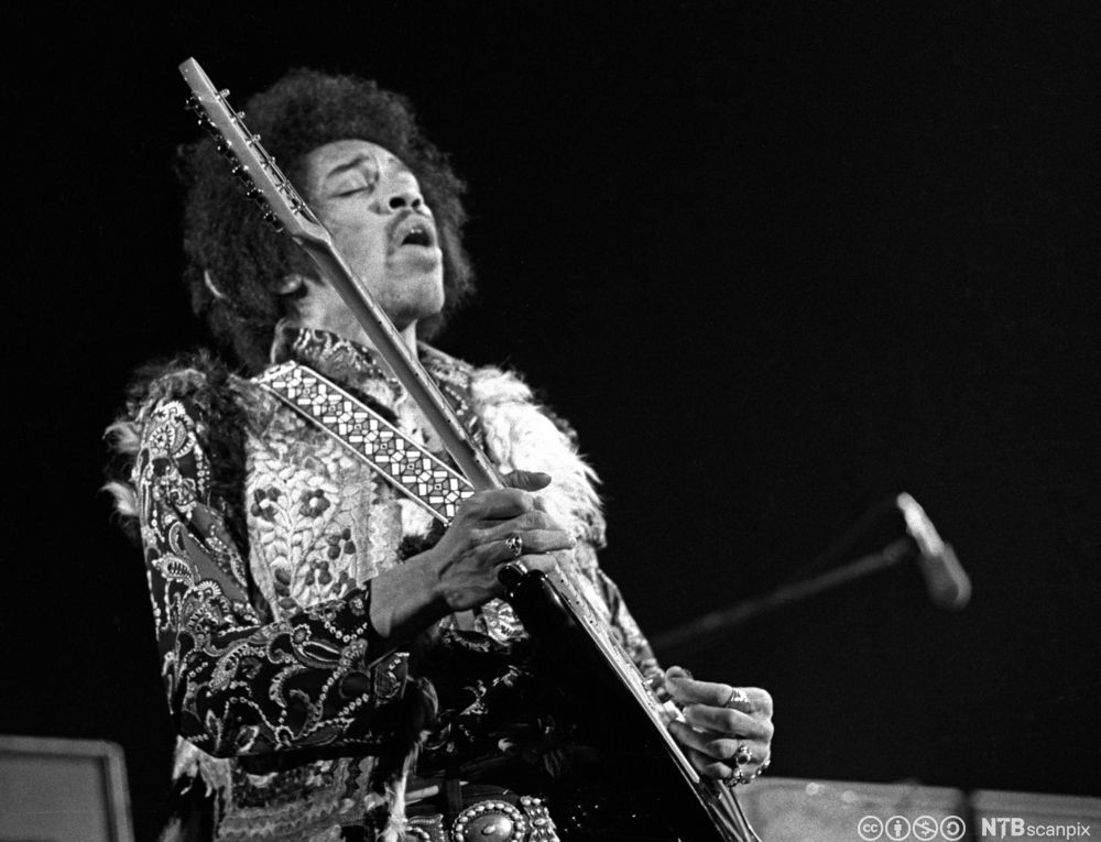 Jimi Hendrix spiller gitar mens han lener seg litt tilbake og lukker øynene. Svart-hvitt-foto.