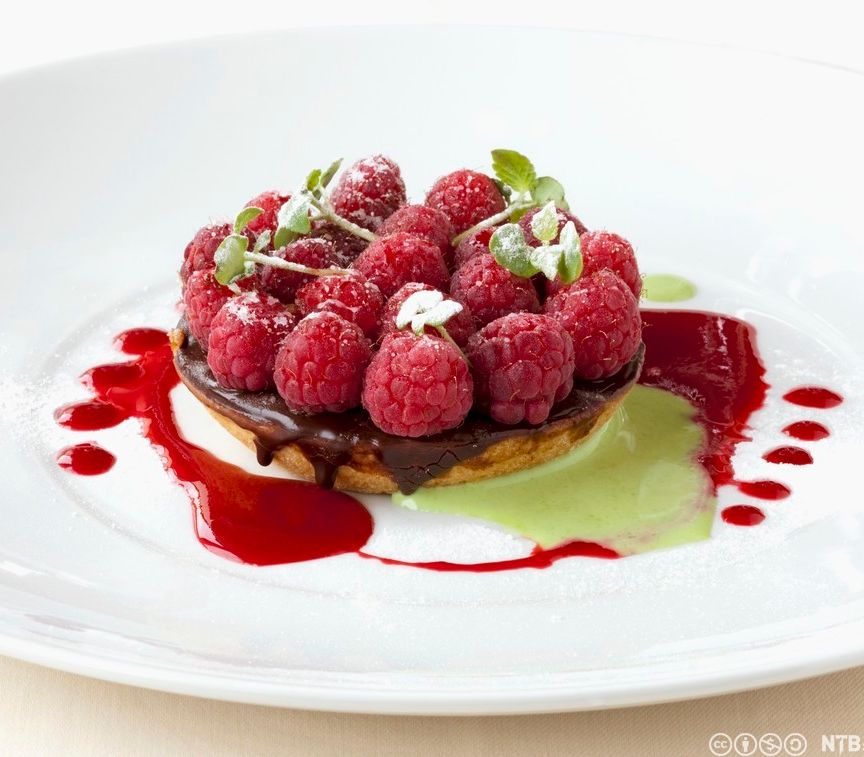 Liten kake med sjokolade dekket av bringebær. Rød og grønn saus rundt på en hvit asjett. Foto.