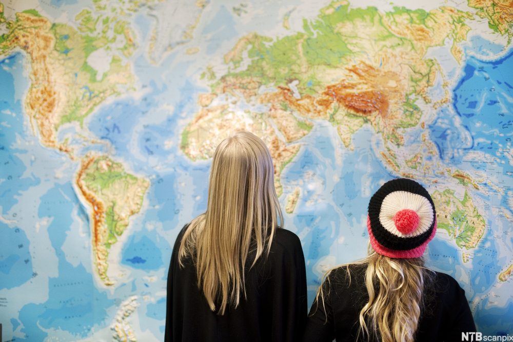 To elever står med ryggen til og betrakter et stort verdenskart på en vegg. Foto.