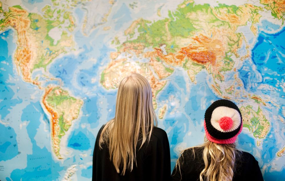 To elever står med ryggen til og betrakter et stort verdenskart på en vegg. Foto.