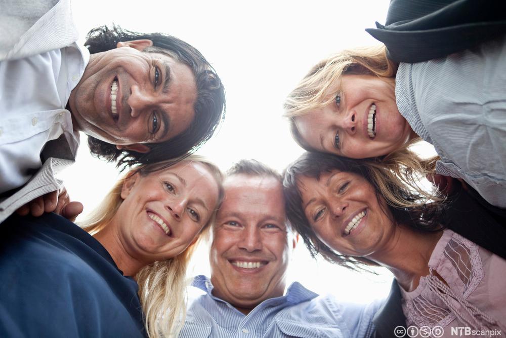 Smilende mennesker står i sirkel, holder om hverandre og kikker ned mot kamera. Foto.