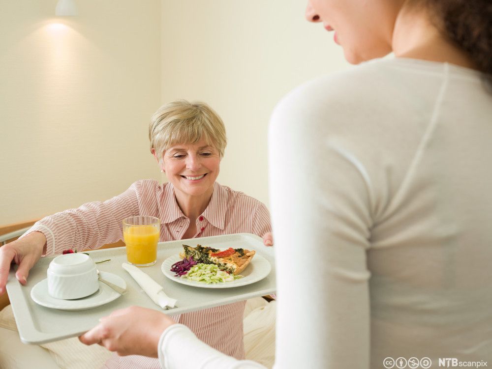 Ei kvinne gir eit brett med mat til ei kvinne som sit i ei seng. Foto.