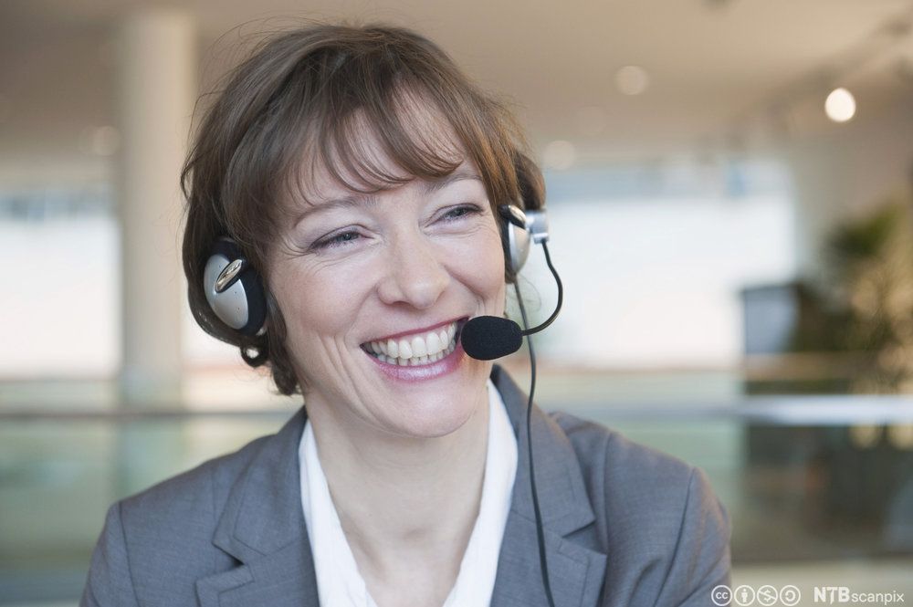 Smilende dame har på seg hodetelefoner med mikrofon. Foto.