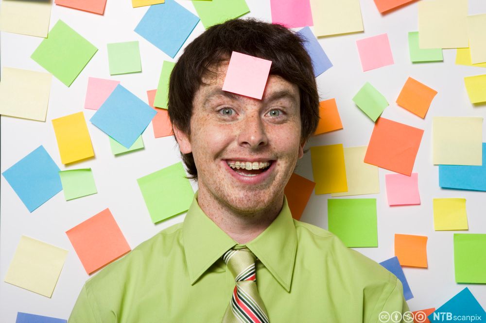 Ung, smilende mann med en post-it-lapp klistret i pannen. Bak ham en vegg full av post-it-lapper i mange farger. Foto.