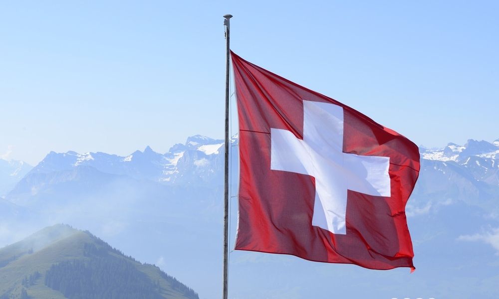 Et kvadratisk rødt flagg med et hvitt kors midt på vaier i vinden. Rigi-fjellet i bakgrunnen. Foto.