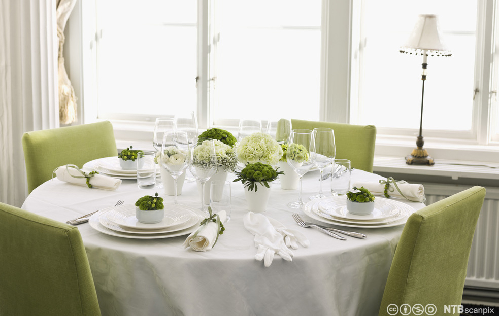 An elegantly set table with a white tablecloth. The chairs are green, as are the decorations on the table. There are white and green flowers. Photo.