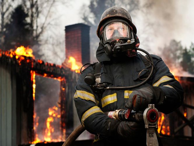 Brannkonstabel med vasslange framfor eit nedbrent hus der restane av huset framleis brenn. Foto.