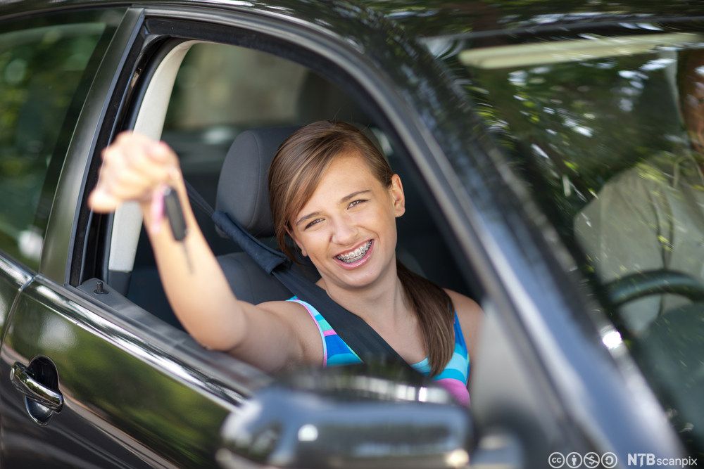 Kvinne sit i ein bil og smiler mens ho held bilnøkkelen opp mot kameraet. Foto.