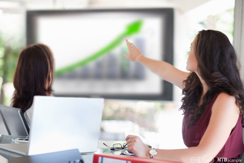 To kvinner ser på en skjerm og peker på et diagram med en grønn pil som peker oppover. Foto.