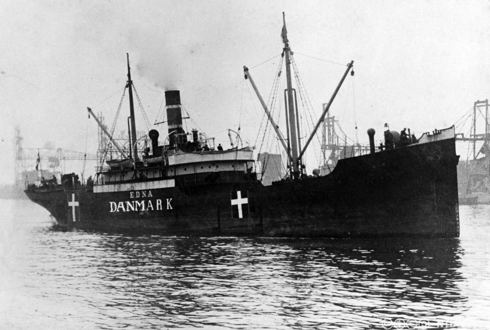 Fotografi av et stort dampskip med navnet Edna og Danmark malt med store bokstaver på siden. To store, danske flagg dominerer også på skipssiden. 