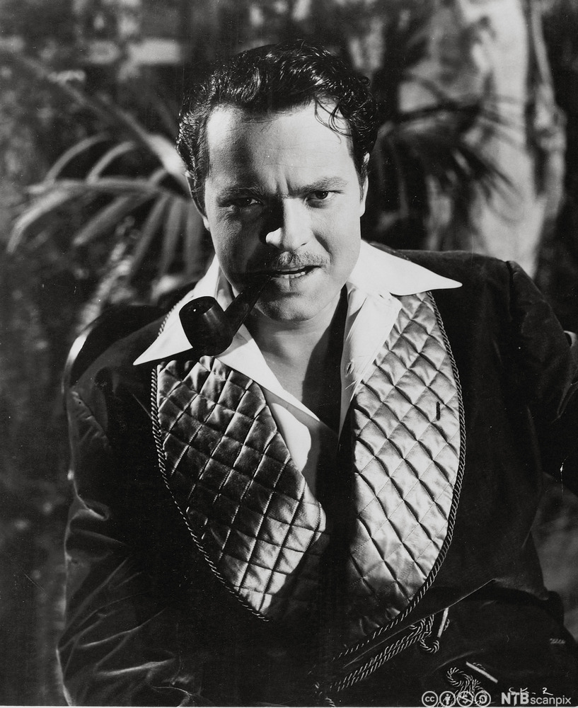 Portrett av skodespelar og regissør Orson Welles. Fotografiet viser ein mann på rundt 40 år med tjukt, relativt kort, svart hår som er greidd bakover. Han har ei pipe i munnen og eit energisk ansiktsuttrykk.
