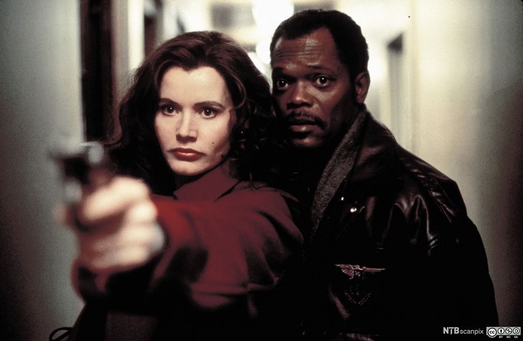 Skuespillerne Geena Davis og Samuel Jackson. Hun sikter med en pistol, og han står tett inntil henne og ser i samme retning. Foto.