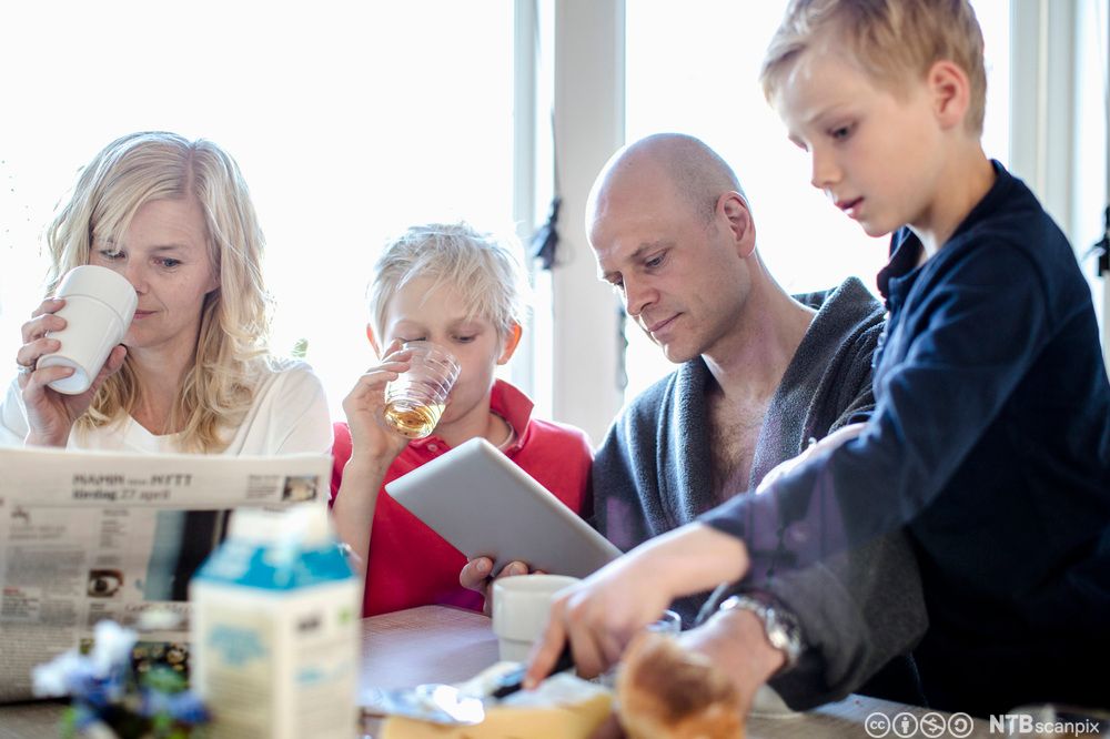 Familie spiser frokost. Familien består av mor, far og to barn. De leser i en papiravis og ser på nettbrett sammen mens de spiser og drikker. Foto.