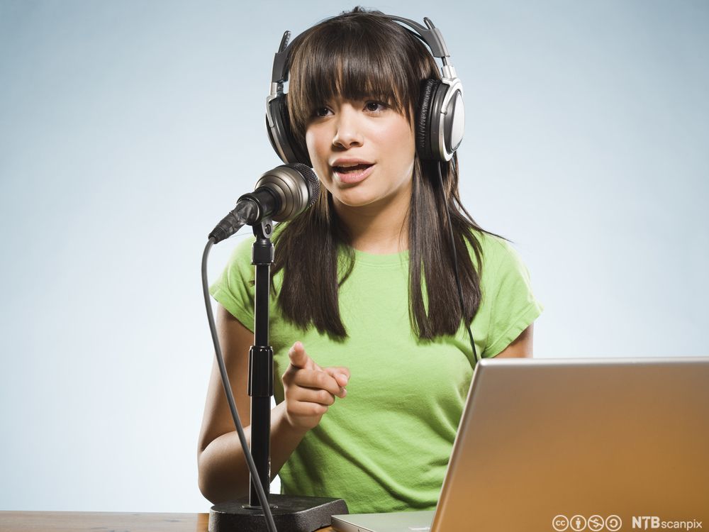 Jente med hodetelefoner og grønn T-skjorte sitter foran en datamaskin og snakker i mikrofon. Foto.