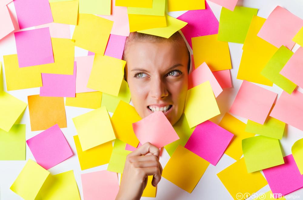 Voksen kvinne dekt til av gule og rosa post-it-lapper. Bare ansiktet og ei hand er synlig. Foto.