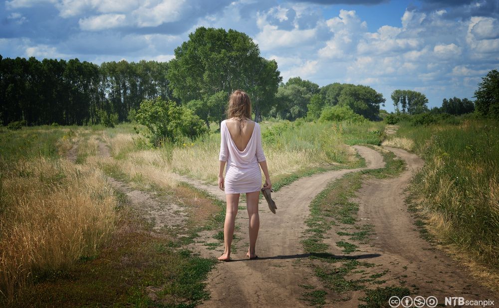 En jente står ved et veiskille med ryggen mot kameraet. Foto.