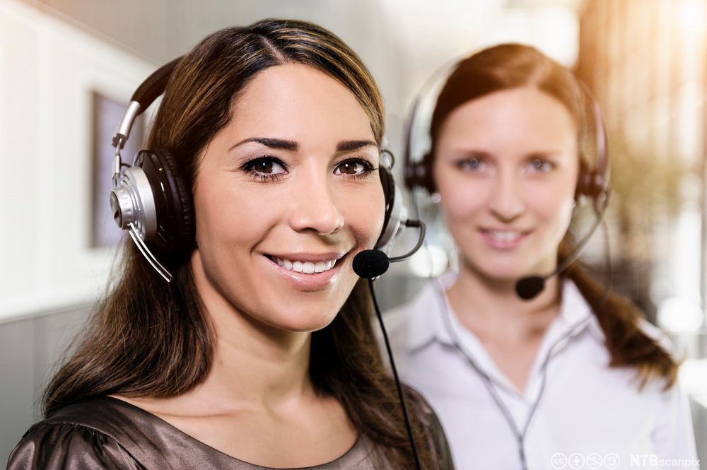 To kvinner med hodetelefon smiler mot kameraet. Foto.