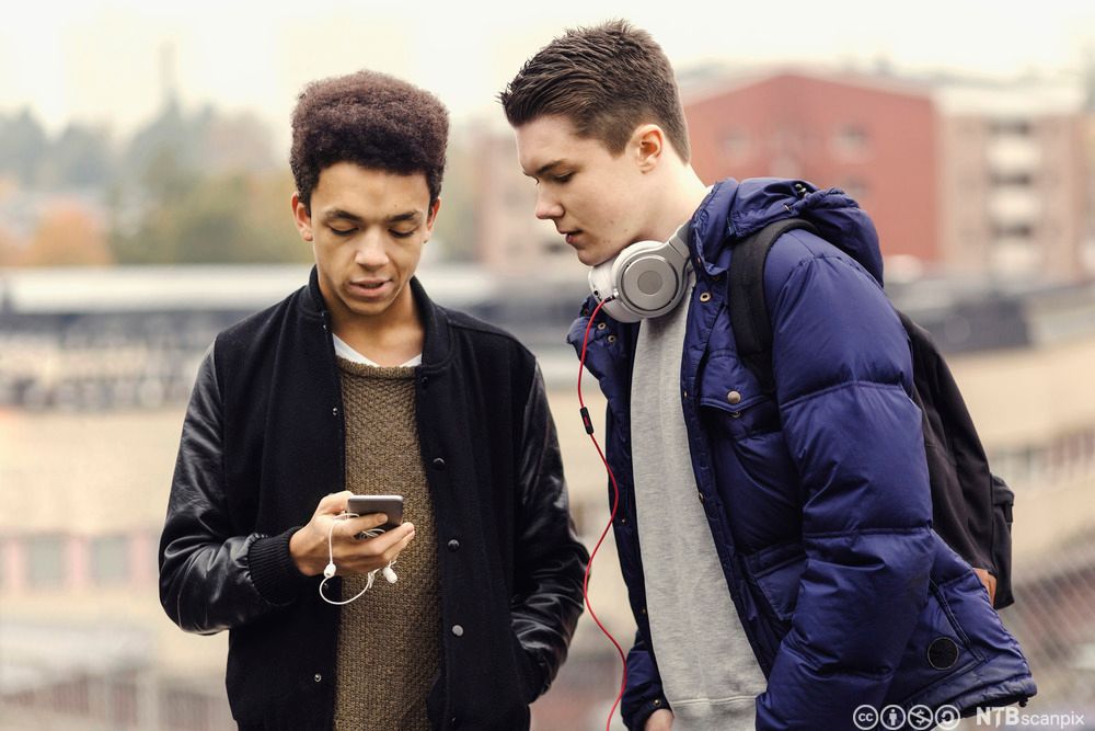 To gutter ser på en mobiltelefon. Den ene gutten bøyer seg fram for å se på mobilen til den andre gutten. Fotografi.