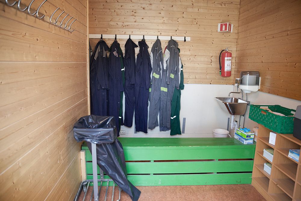 Sluse med hygiene- og smittevernutstyr som vask, plastovertrekk til sko, kjeledresser og søppelpose. En benk på tvers av rommet skiller mellom rein og urein side. Foto.