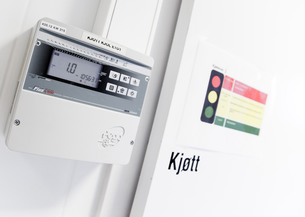 Dør til kjølerom for kjøtt. Ved siden av døra henger et panel som viser temperatur og klokkeslett og har seks knapper for ulike innstillinger. På døra er det klistret opp et trafikklyssymbol i rødt, gult og grønt med noen instrukser. Foto.