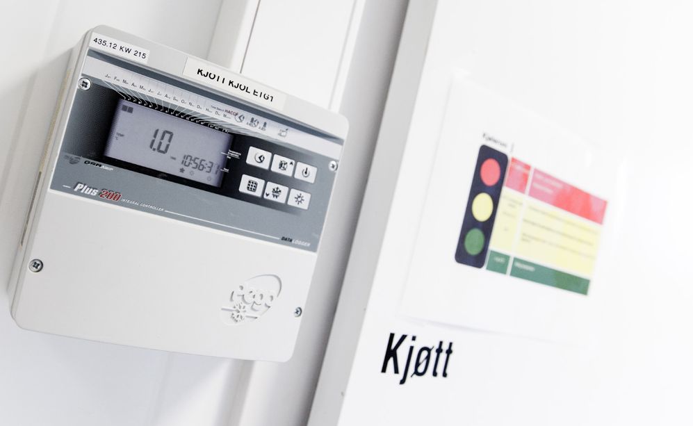 Dør til kjølerom for kjøtt. Ved siden av døra henger et panel som viser temperatur og klokkeslett og har seks knapper for ulike innstillinger. På døra er det klistret opp et trafikklyssymbol i rødt, gult og grønt med noen instrukser. Foto.