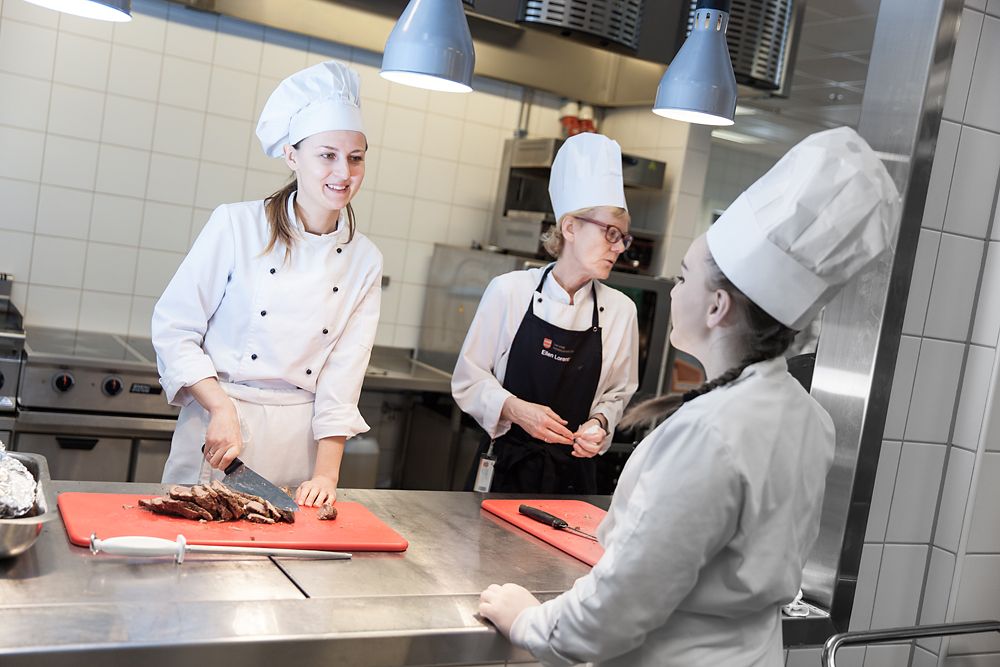 Tre kvinnelege kokkar i samtale på kjøkkenet. Ei av dei skjer opp kjøt. Foto.