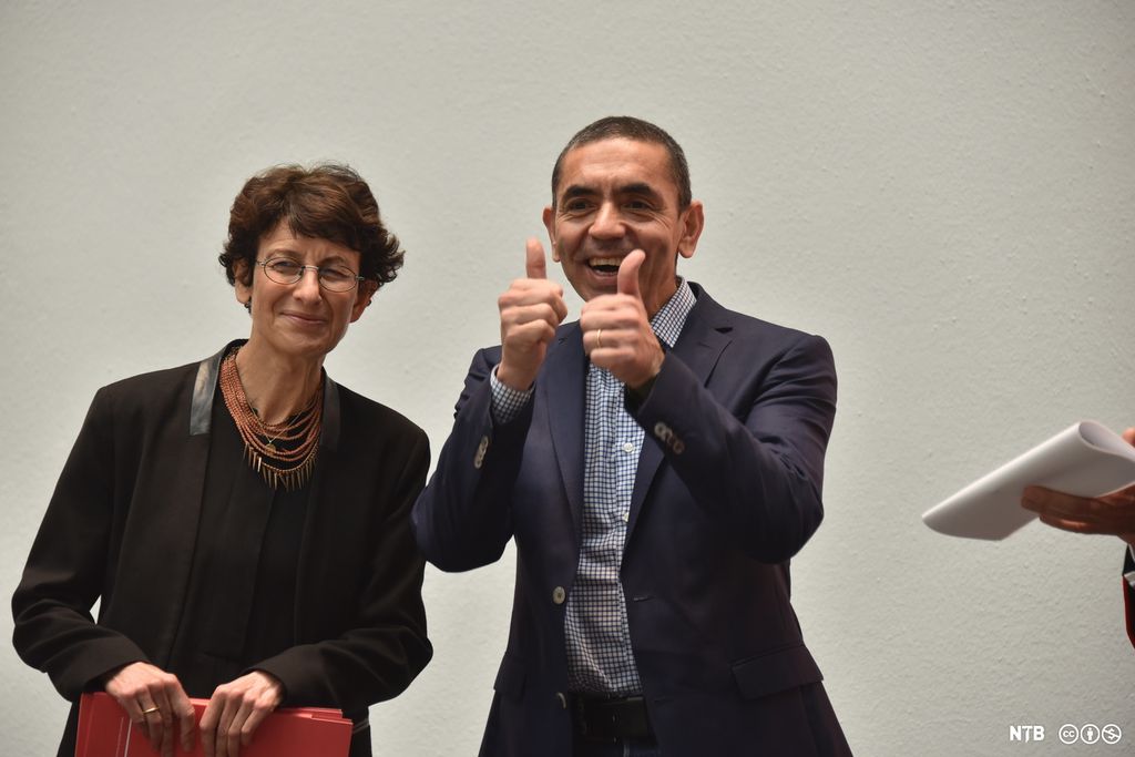 En leende mann i mørkeblå dress og blå-og-hvitt-rutete skjorte som viser tommel-opp-tegnet med begge hendene. Ved siden av ham en smilende kvinne kledd i sort bluse og jakke. Hun har et smykke rundt halsen som består av mange tynne rader med korallfargede perler. Foto.