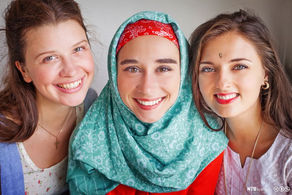 Tre unge jenter: ei med kors i halsen, ei med hijab på hodet og ei med bindi i pannen. Jentene smiler mot kamera. Foto.