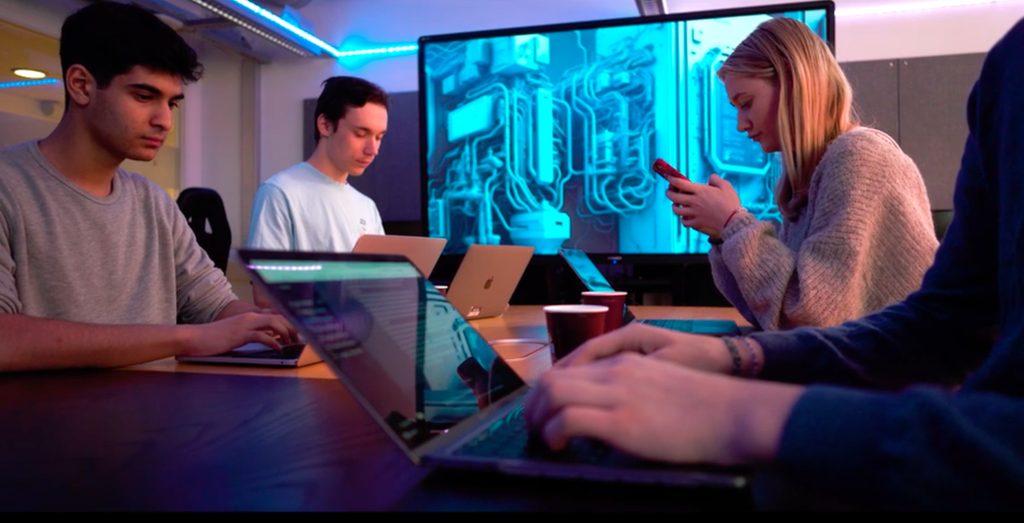 Fire ungdommer sitter rundt et bord og jobber med datamaskiner og mobiltelefoner. Foto.