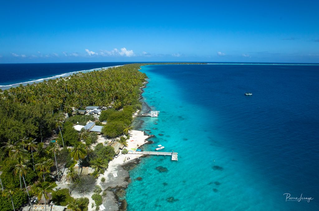 Lang og smal øy til venstre i bildet, med skog, noen få hus og ei brygge. Vannet er klart, og havbunnen skinner igjennom der vannet er grunt. Foto.