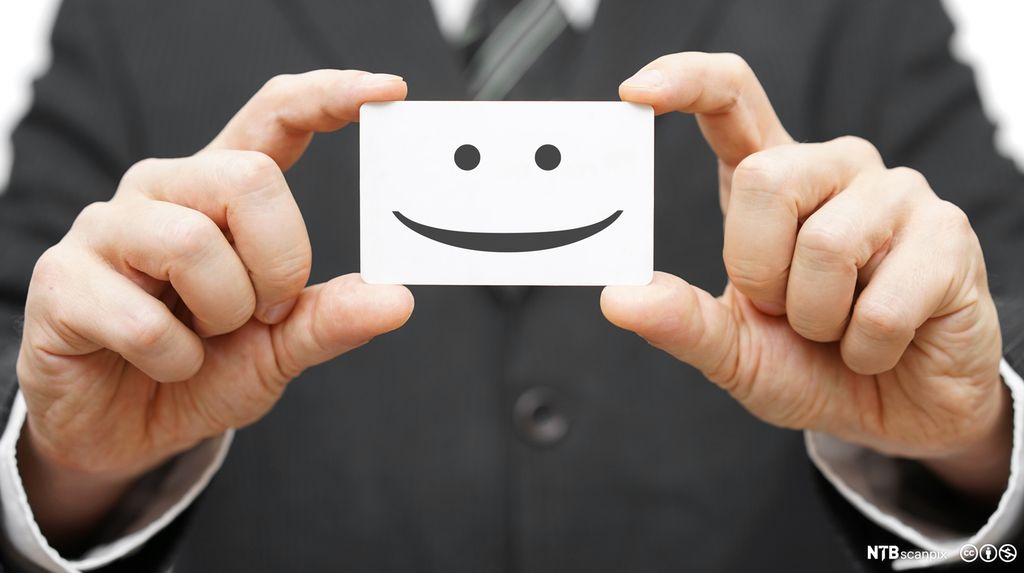 En mann i svart dress holder opp et hvitt kort med smilefjes på. Foto.