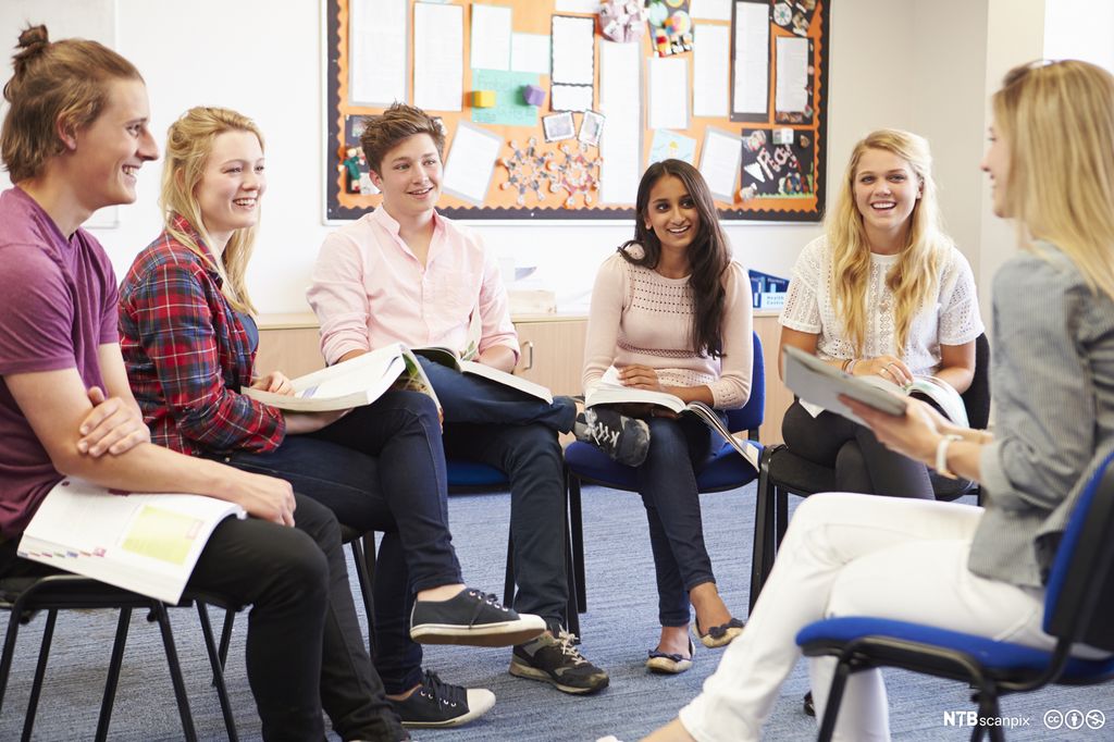 Seks ungdommer sitter i ring i et klasserom og diskuterer fag. De smiler og ser på hverandre. Foto.