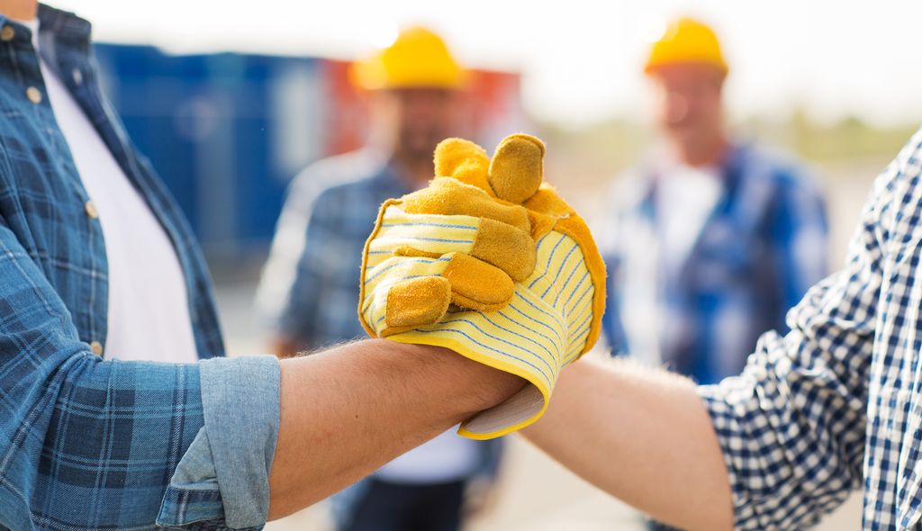 To personer med arbeidshansker håndhilser. To menn med gule hjelmer står i bakgrunnen. Foto.