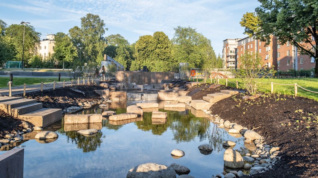 Bekk med steinar og trappetrinn i ein park, omgitt av jord, tre og bustadblokker i bakgrunnen. Foto.