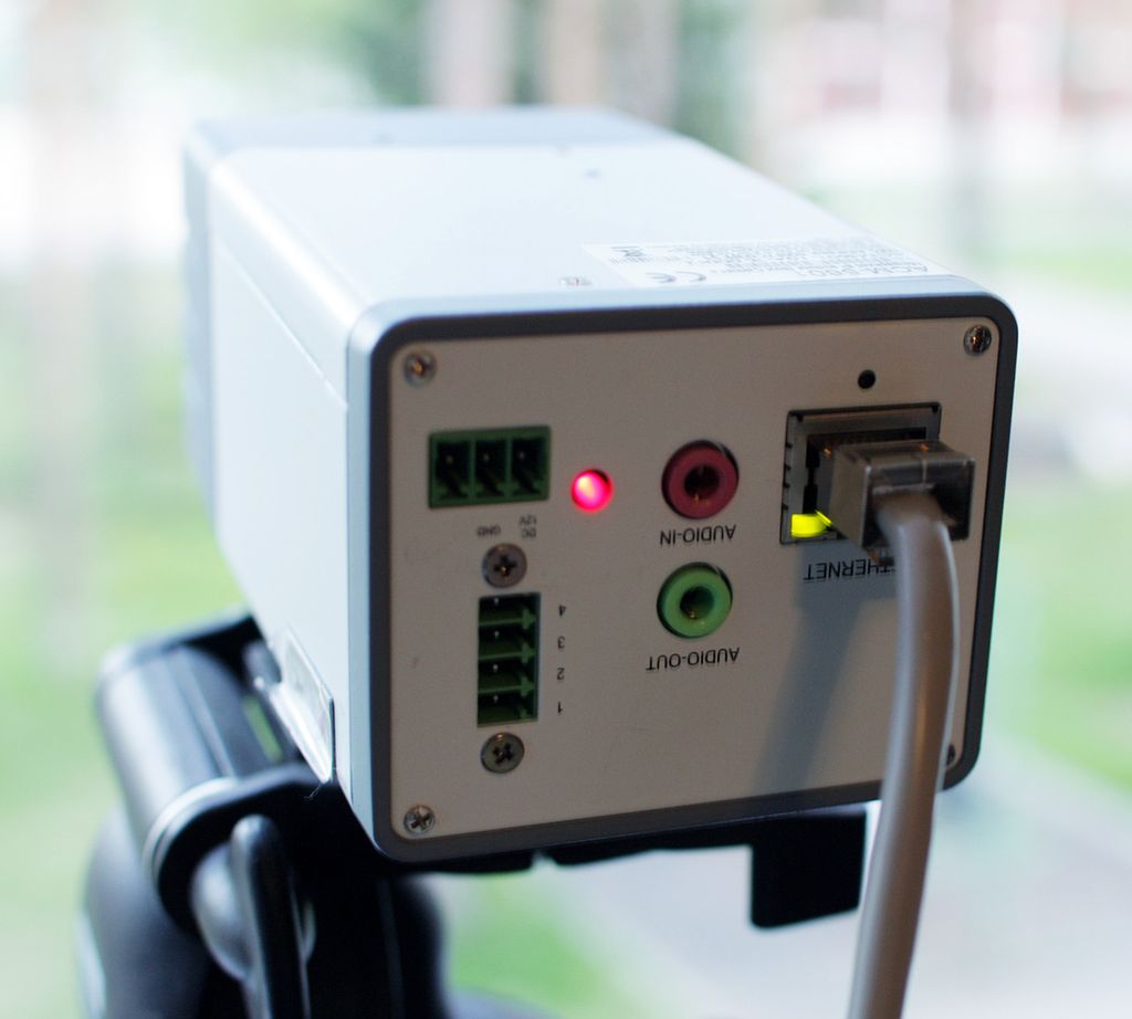 Baksida av eit lite firkanta kamera. Eit lys viser at kameraet er på. Berre ein nettverkskabel er tilkopla. Foto